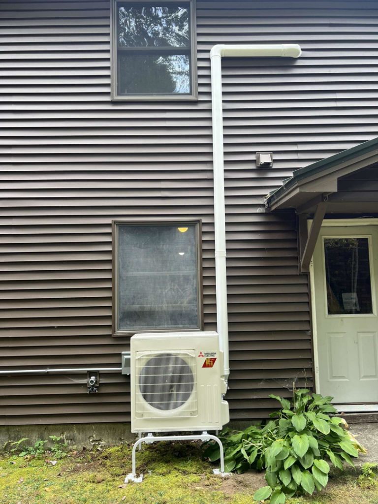 An elevated Mitsubishi heat pump outdoor unit installed on the side of a brown house by S&J Heat Pumps in Augusta, ME.