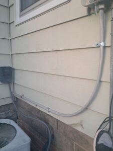 Exterior electrical conduit running along a house wall, connecting to an AC unit, installed by Titus & Son's Electric in Bluffton, IN.