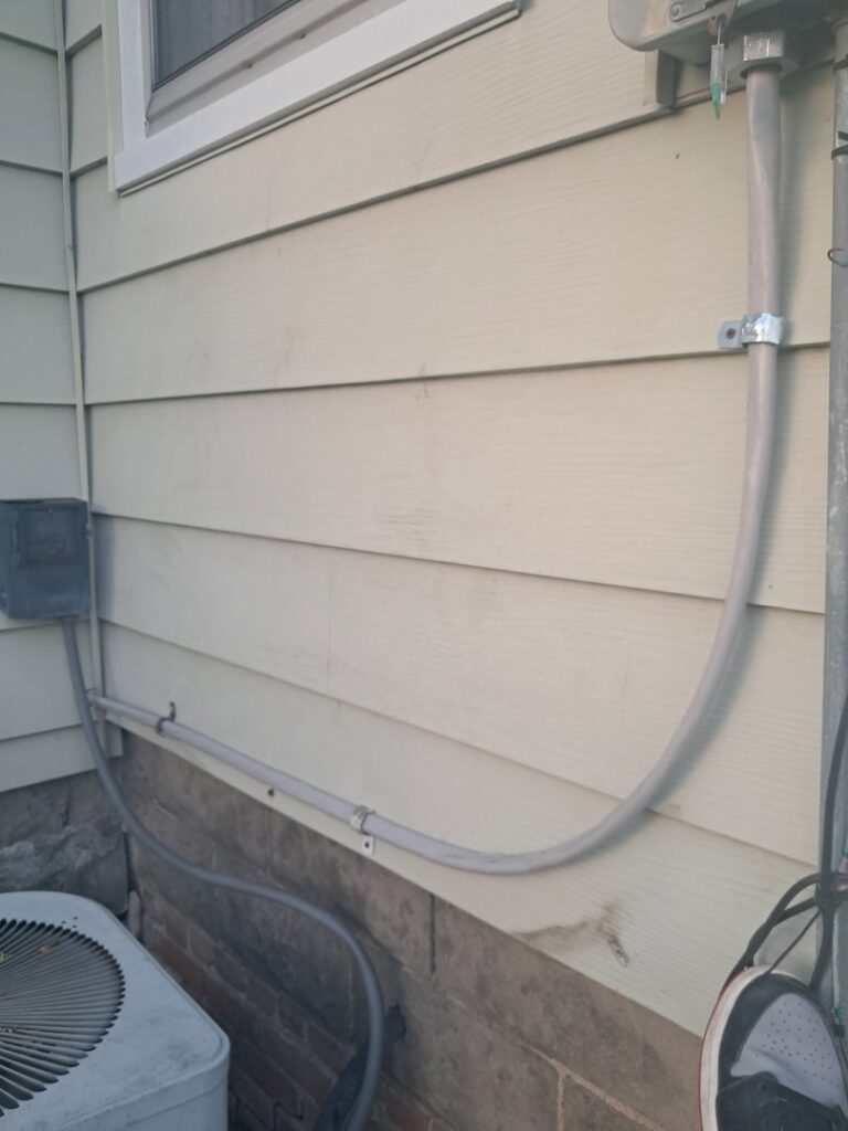 Exterior electrical conduit running along a house wall, connecting to an AC unit, installed by Titus & Son's Electric in Bluffton, IN.