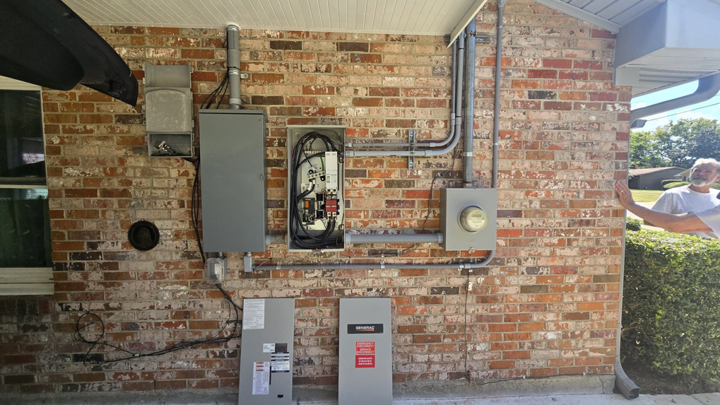An exterior view of electrical panels, conduits, and a generator transfer switch installation on a brick wall by Mcgehee Electric, LLC in Montgomery, AL.