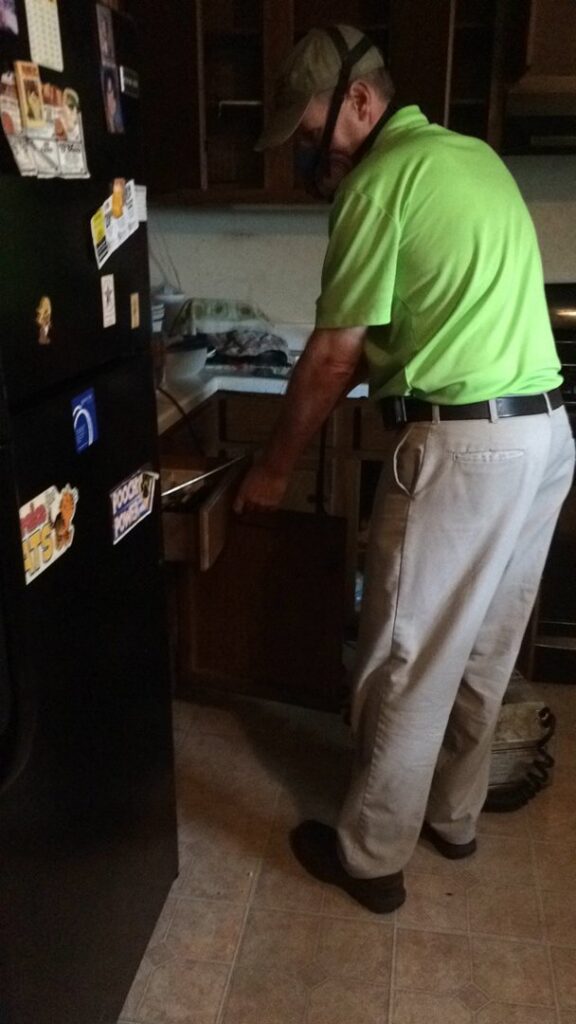 An exterminator wearing a respirator, inspecting under a kitchen cabinet for pests at a client's home in Roanoke, VA, by Perdue Exterminators.