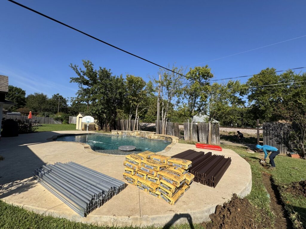 Fencing materials, including metal posts and concrete bags, with a worker digging a trench for installation by Dallas Fence in Dallas, TX.