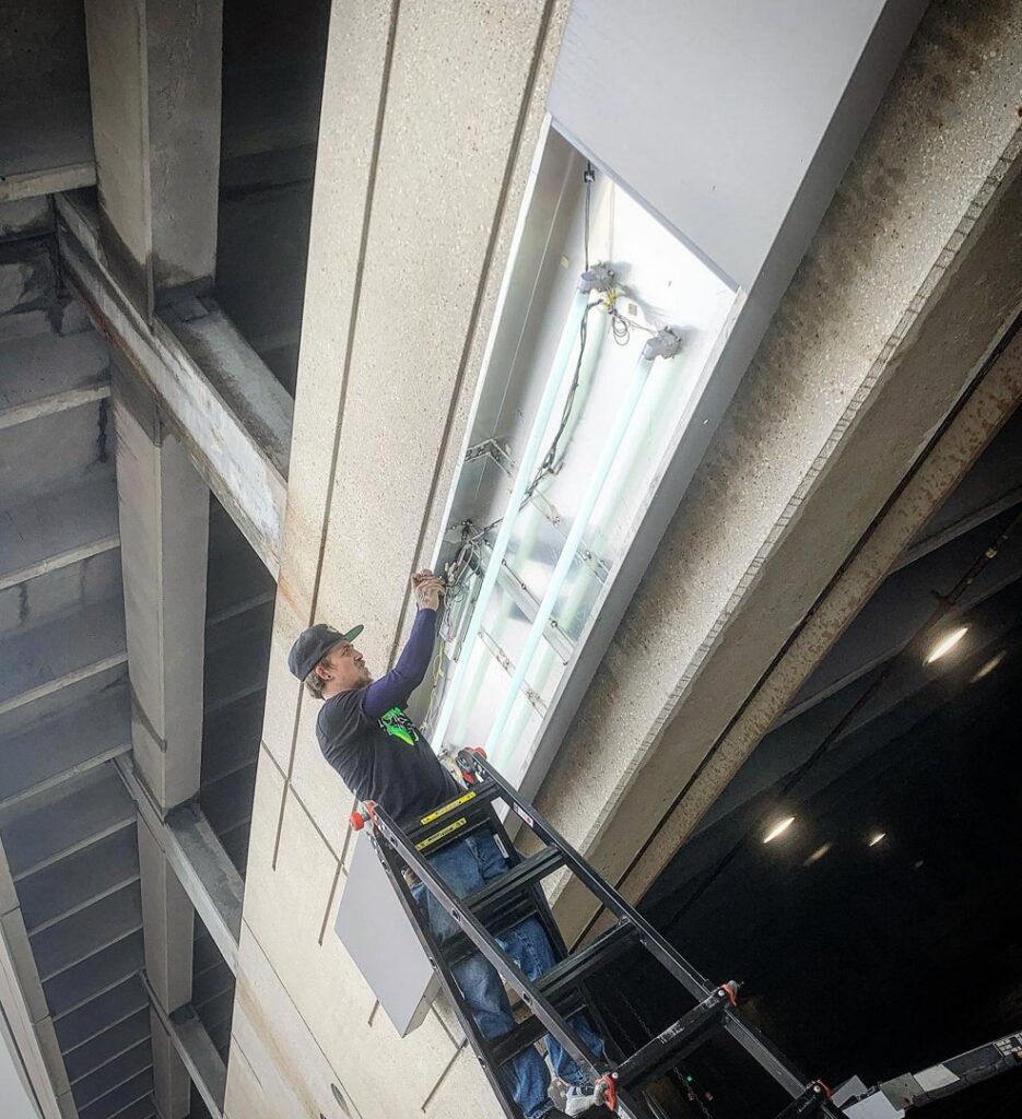 An electrician from Lumenus Technologies, LLC repairing a fluorescent light fixture in Lenexa, KS.