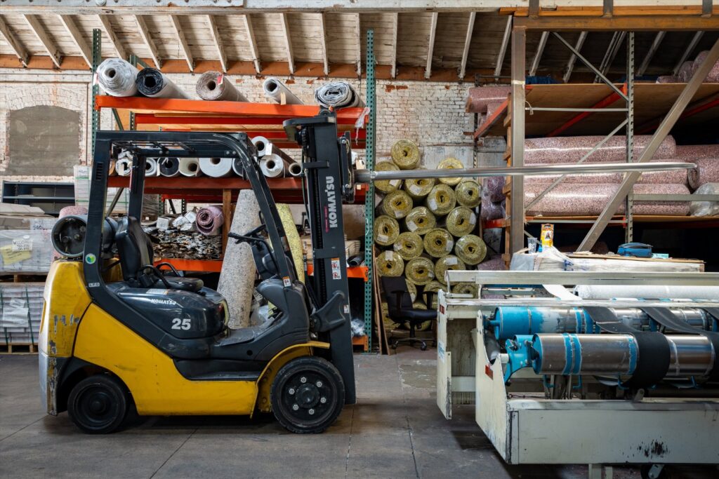A forklift moving a large roll of carpet onto a cutting machine at Mill Sales Co. in Los Angeles, CA