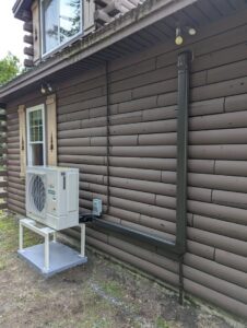 A Fujitsu heat pump outdoor unit installed on the side of a log cabin style house by Kinney Family Heat Pumps in Augusta, ME.