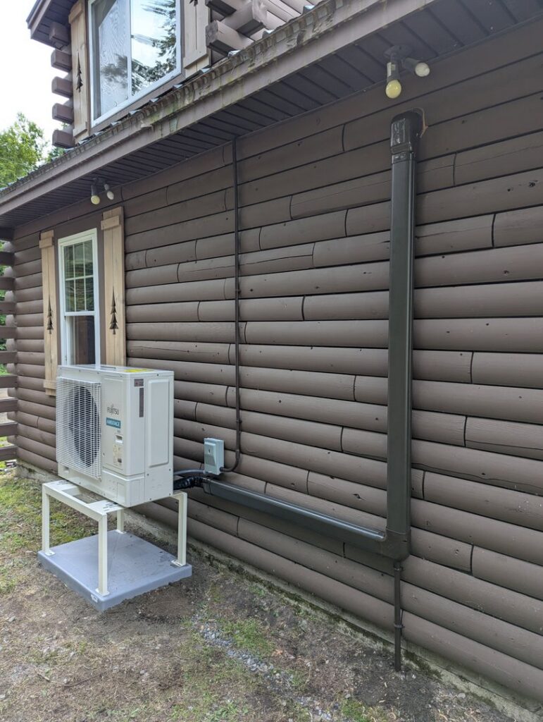 A Fujitsu heat pump outdoor unit installed on the side of a log cabin style house by Kinney Family Heat Pumps in Augusta, ME.