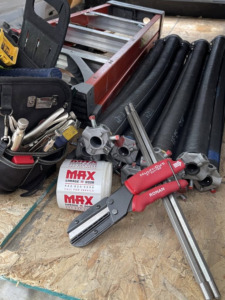 Various garage door repair tools, springs, and Max Garage Door stickers laid out for a job in Minneapolis, MN.