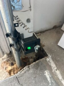 A close-up of a garage door safety sensor with a green indicator light, installed by BSD Garage Door Service in Naperville, IL.