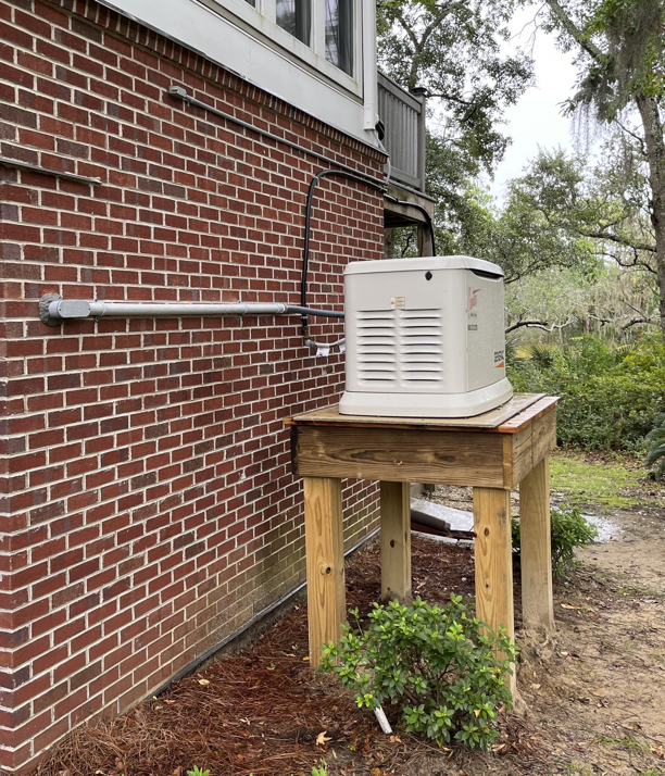 A Generac generator installed on a wooden stand next to a brick house by Strother & Son Electric in Mount Pleasant, SC.