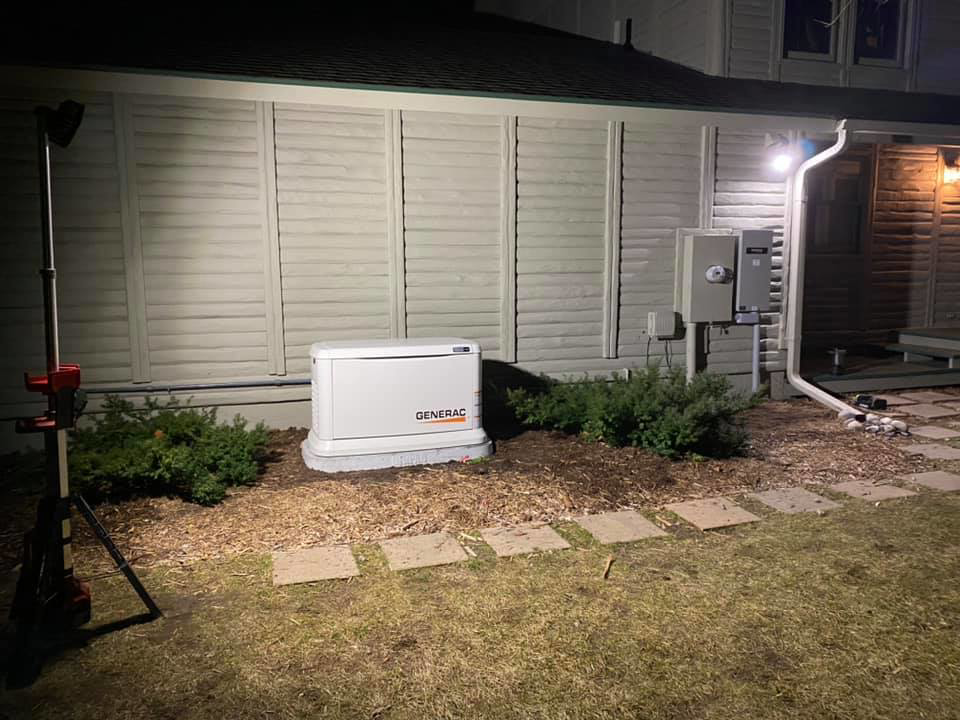 A Generac generator installed next to a house at night, illuminated by a work light, by Great Lakes Power Generation in Williamsburg, MI.