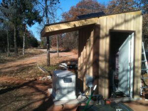 A generator installed next to a small shed in a wooded area with visible electrical conduit by Generator Experts of Oklahoma in Norman, OK.
