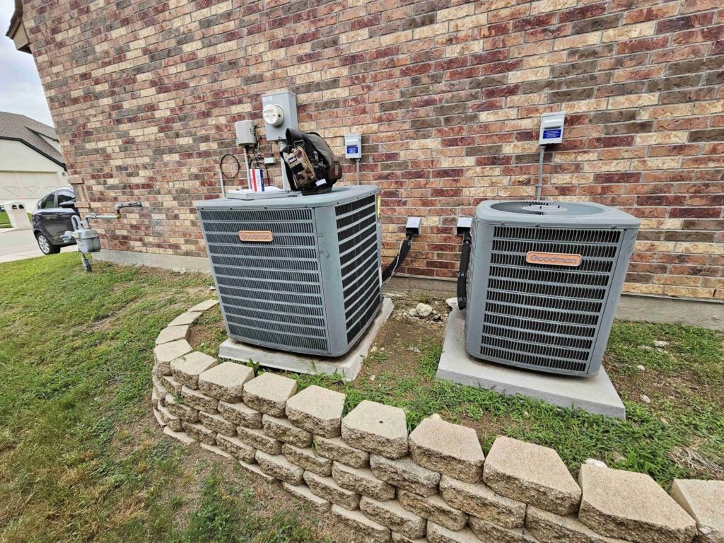 Two Goodman outdoor AC units next to a brick house, with one unit appearing to be under service by Fort Worth Air Conditioning Co. Inc. in Fort Worth, TX.