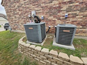 Two Goodman outdoor AC units next to a brick house, with one unit appearing to be under service by Fort Worth Air Conditioning Co. Inc. in Fort Worth, TX.