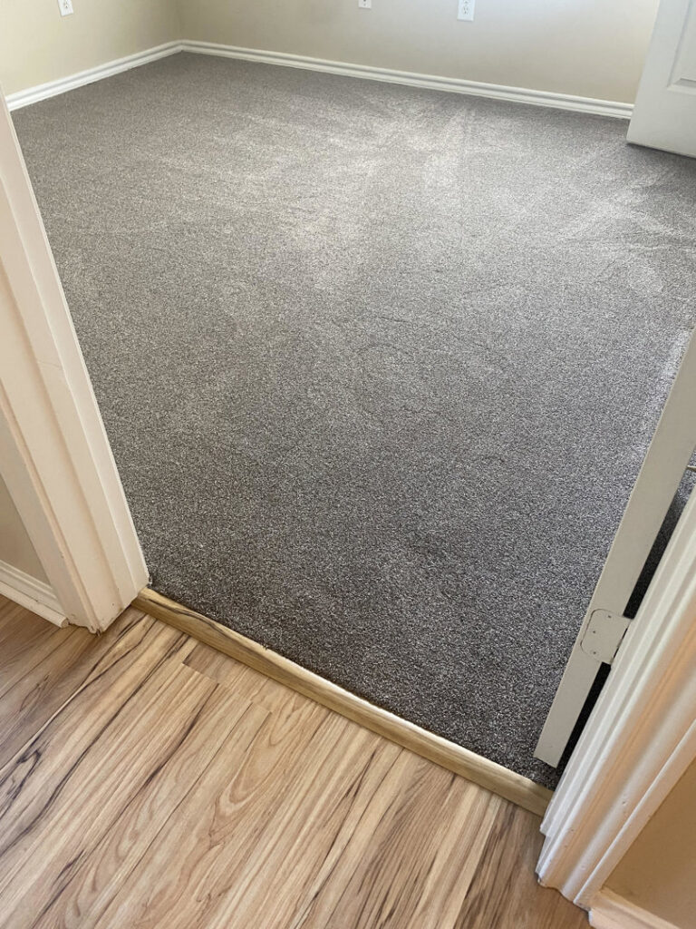 Newly installed gray carpet in a room, transitioning to wood-look flooring by Floors For Living - Meyerland in Houston, TX.