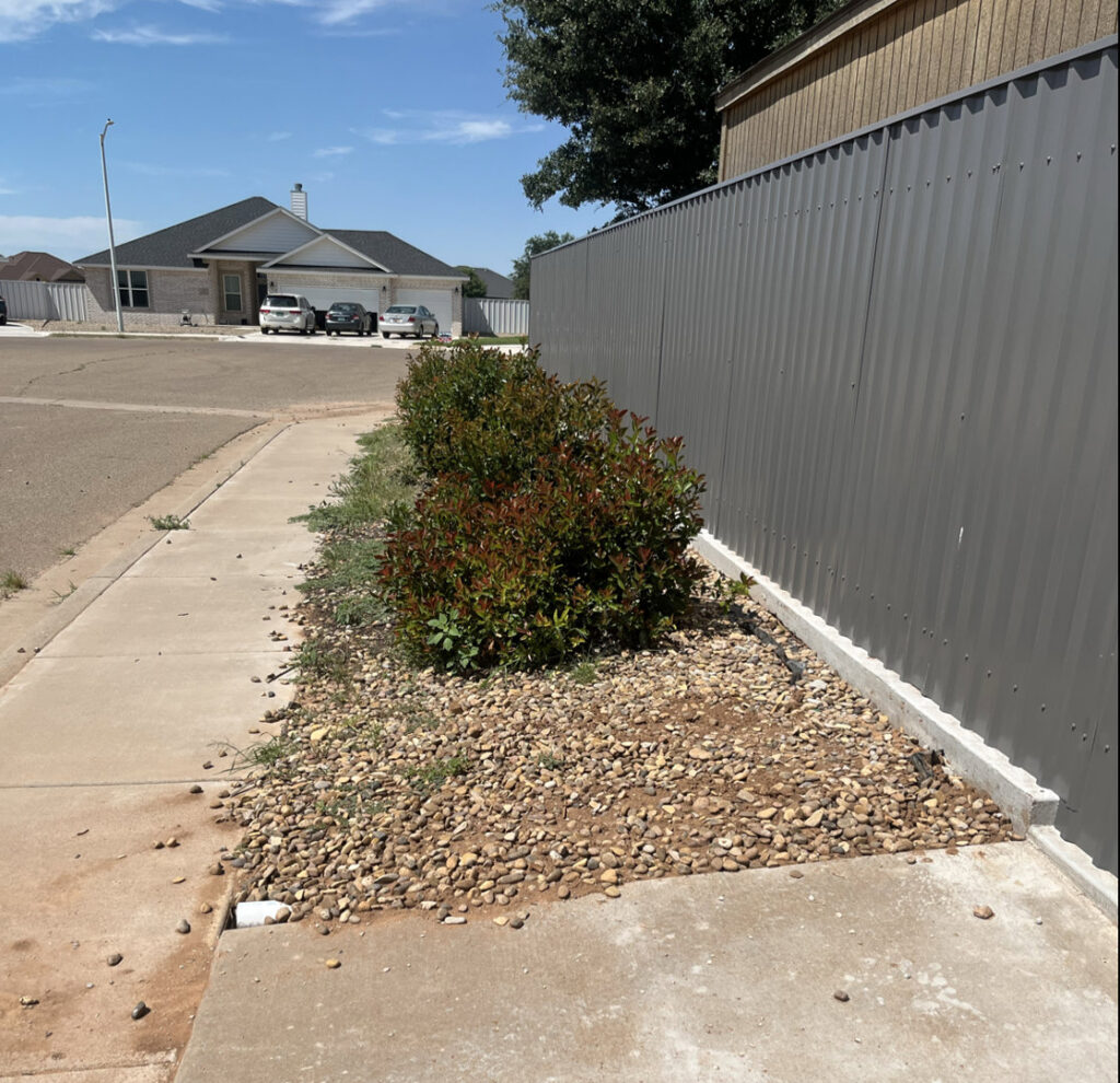 A modern gray corrugated metal fence installed along a sidewalk with landscaping, completed by DC Fencing LLC in Clovis, NM.