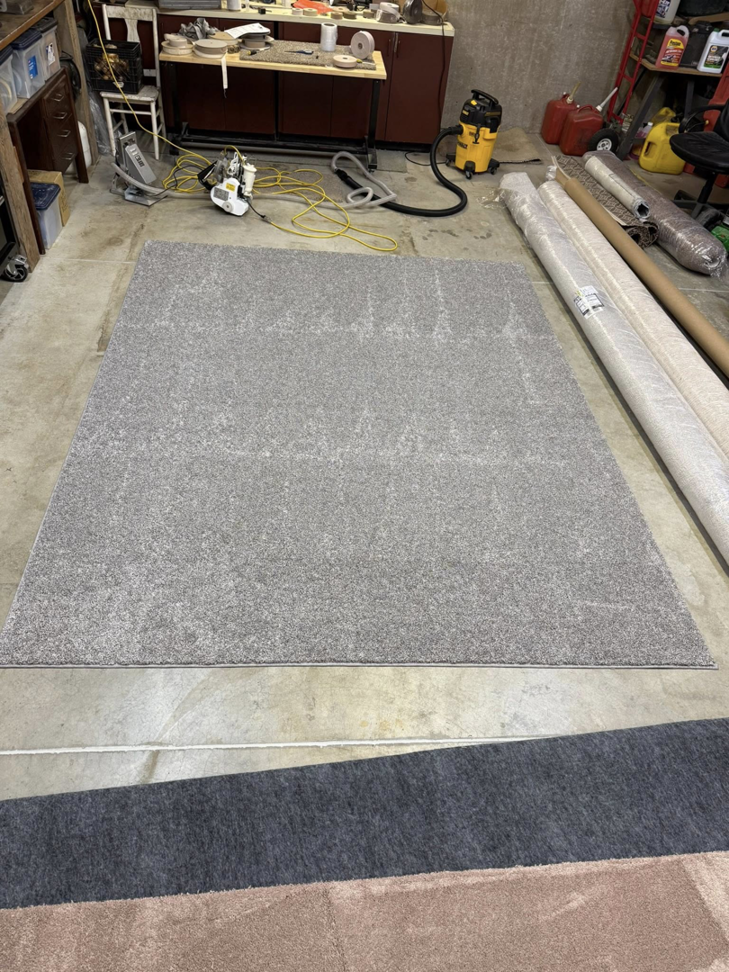 A grey area rug laid out with rolls of carpet material in the background, at Rug Edge Binding Company in Spokane, WA