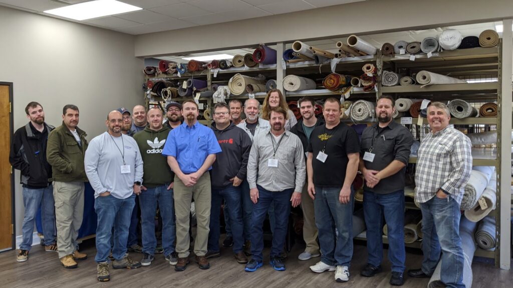 A group of men standing in a rug storage facility with many rolled rugs, at the Association of Rug Care Specialists in Salem, OR