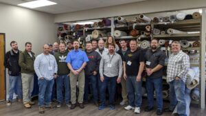 A group of men standing in a rug storage facility with many rolled rugs, at the Association of Rug Care Specialists in Salem, OR