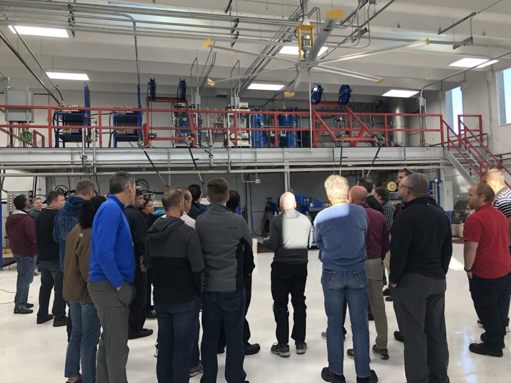 A group of professionals touring a rug care facility, observing machinery at the Association of Rug Care Specialists in Salem, OR