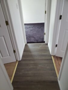 A hallway with wood flooring transitioning into a room with dark grey carpet by The Floor Store Inc. in Scranton, PA.