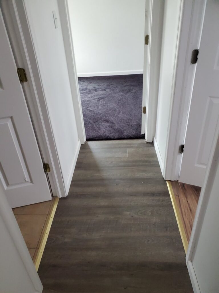 A hallway with wood flooring transitioning into a room with dark grey carpet by The Floor Store Inc. in Scranton, PA.