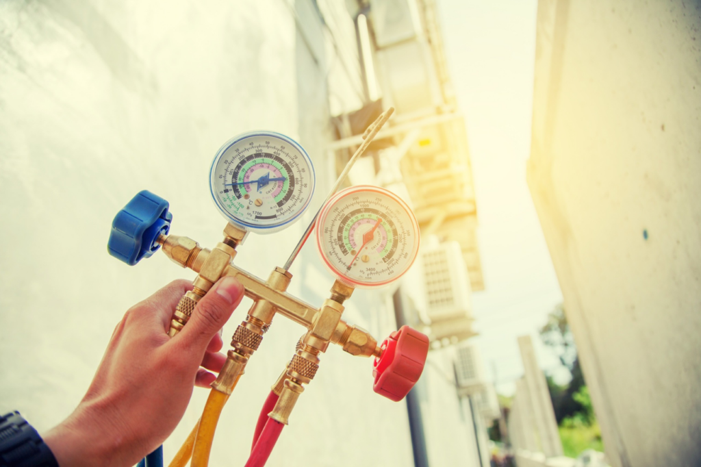 A hand holding HVAC manifold gauges with outdoor air conditioning units in the background for Chrismon Heating & Cooling in Greensboro, NC.