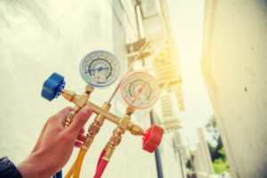A hand holding HVAC manifold gauges with outdoor air conditioning units in the background for Chrismon Heating & Cooling in Greensboro, NC.