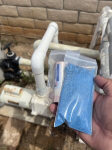 A hand holding bags of blue granular pool chemicals, used for treatment by Good Guy Pool Service in Moreno Valley, CA.