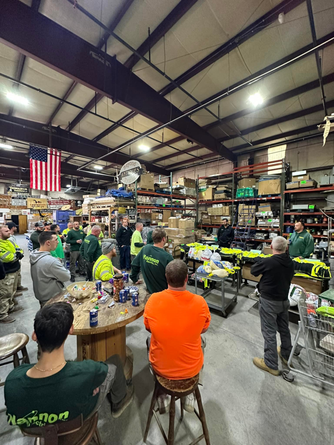 The Hannon Electric Inc. team gathered for a meeting in their South Easton, MA warehouse, surrounded by electrical supplies.