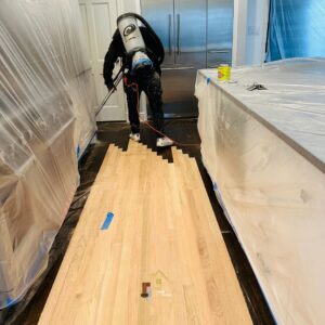 A worker applying finish or sanding a hardwood floor, with the kitchen area protected, by Isad Home Floor in Bridgeport, CT.