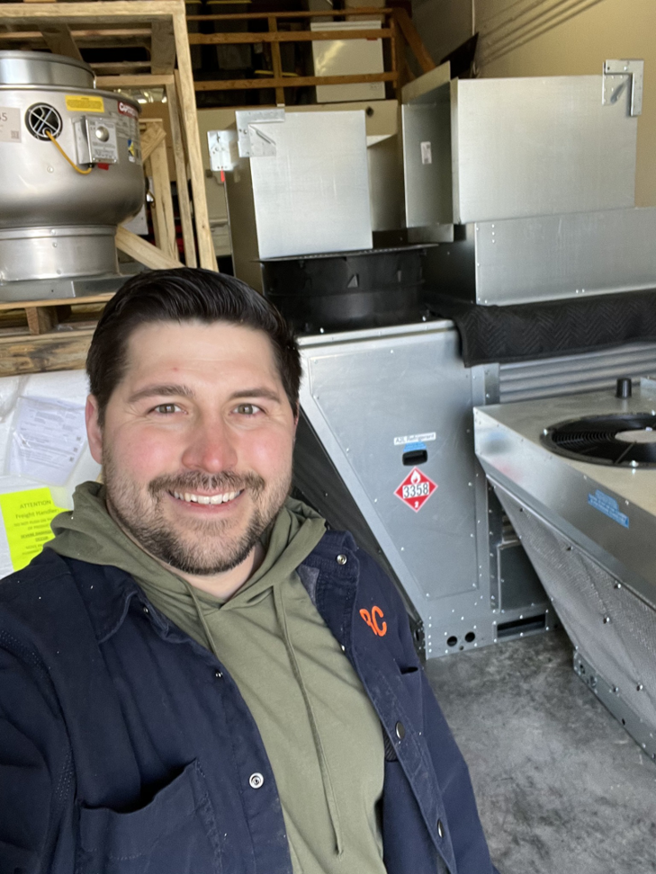 An HBC HVAC technician smiling next to new heating and cooling equipment ready for installation in Cranston, RI.