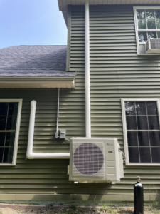 A heat pump outdoor unit installed on the green siding of a residential home by S&J Heat Pumps in Augusta, ME.