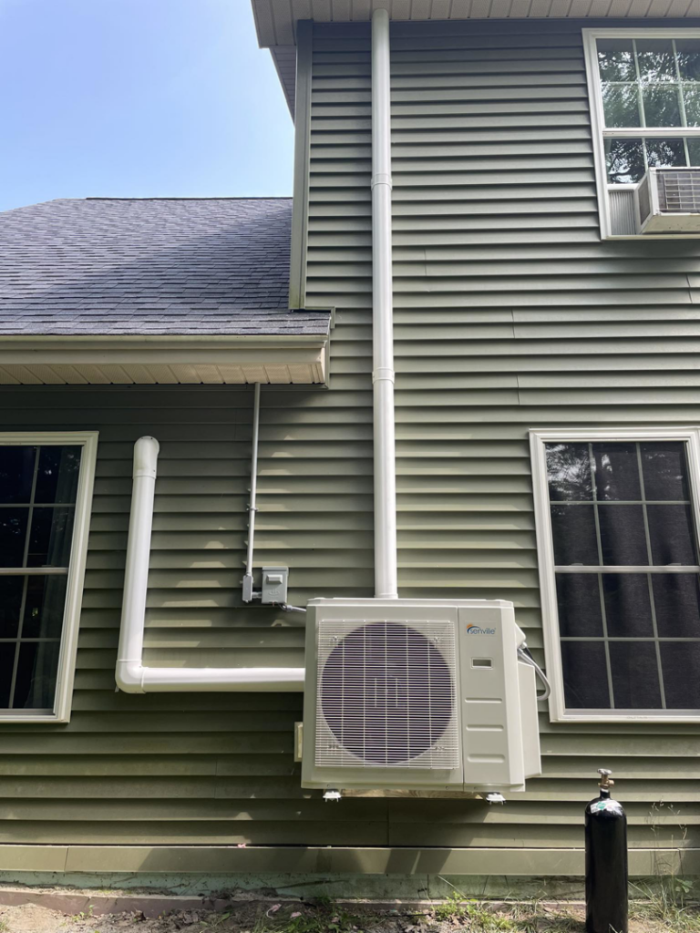 A heat pump outdoor unit installed on the green siding of a residential home by S&J Heat Pumps in Augusta, ME.