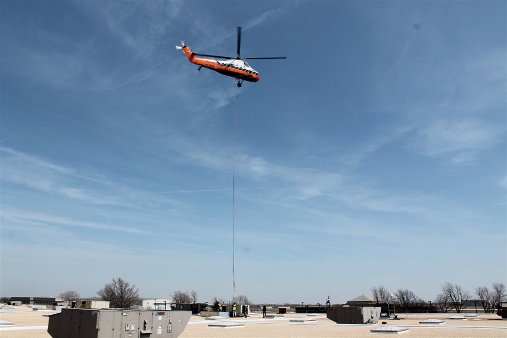 A helicopter lifting a large rooftop HVAC unit for installation or replacement by DMC Service, Inc. in Olathe, KS.