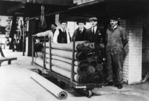 A historical photo of a delivery crew with rolled carpets on a cart at American Rug Laundry in Saint Paul, MN.