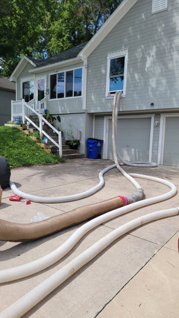 Hoses extending into a house window, likely for attic insulation or pest treatment by KC Pest Experts in Olathe, KS.