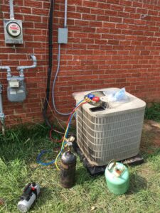An HVAC technician performing refrigerant service on an outdoor AC condenser unit with gauges and tanks by The A/C Guy in Shreveport, LA.