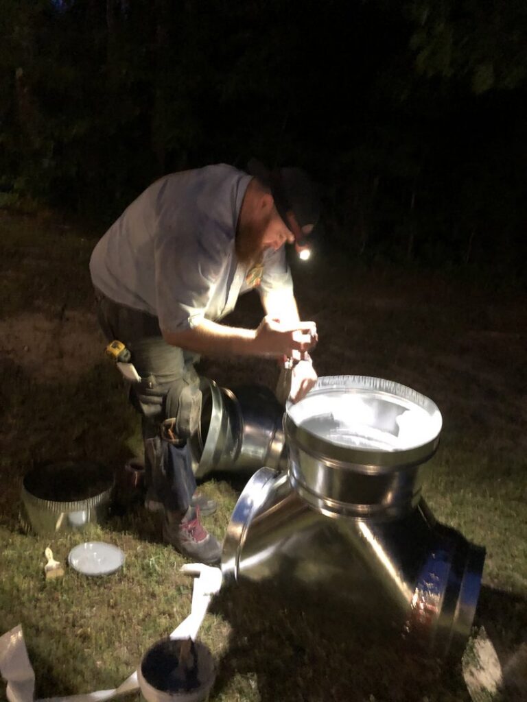 An HVAC technician assembling metal ductwork components outdoors at night for Workman Mechanical in Graham, NC.