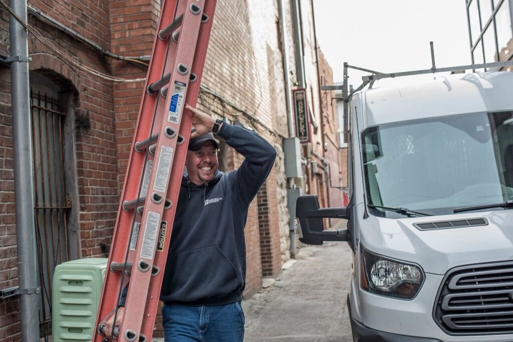 A smiling HVAC technician carrying a ladder next to a service van for QuesTec in Columbia, MO