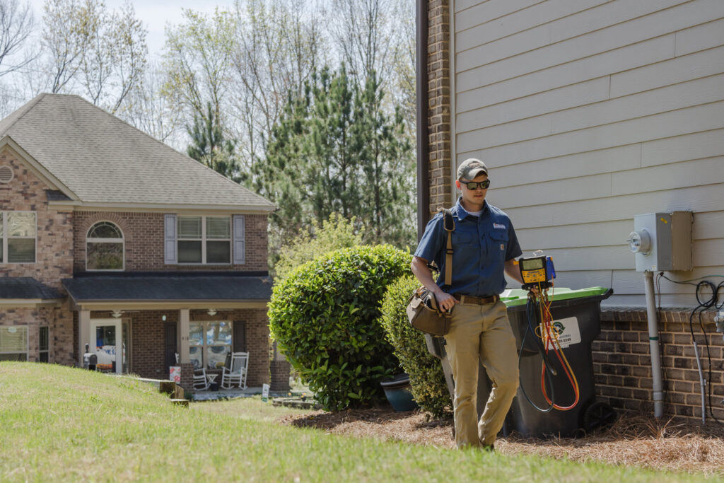An HVAC technician carrying tools near an outdoor AC unit at a residential job for On Call Plumbing, Heating & Air in Columbia, SC