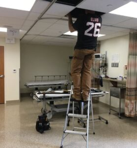 A HVAC technician on a ladder performing ceiling repair in a commercial setting for Tom's Heating & Air Conditioning LLC in Lacey Township, NJ.