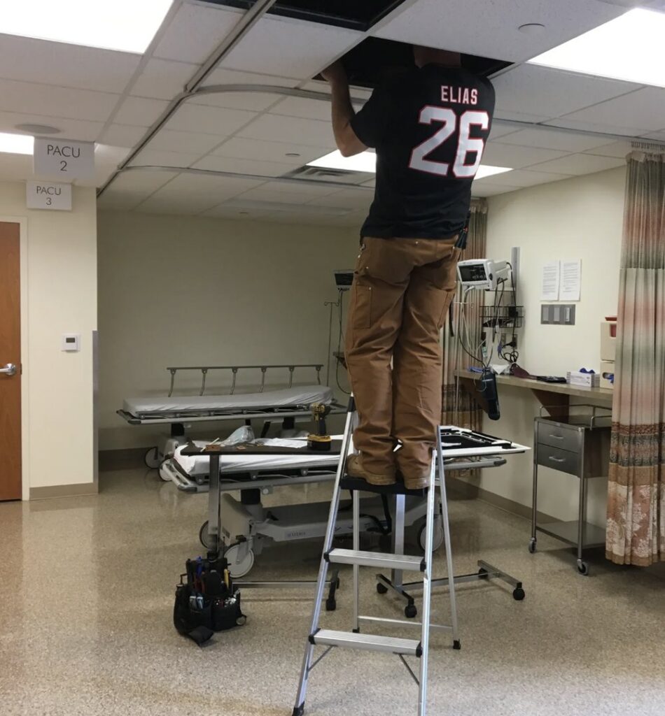 A HVAC technician on a ladder performing ceiling repair in a commercial setting for Tom's Heating & Air Conditioning LLC in Lacey Township, NJ.