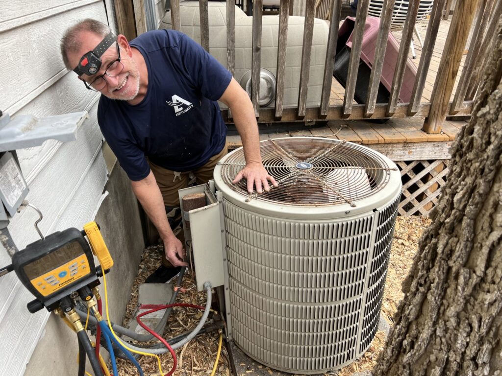 A smiling HVAC technician checking an outdoor AC unit with gauges for Airtegrity Comfort Solutions in San Antonio, TX