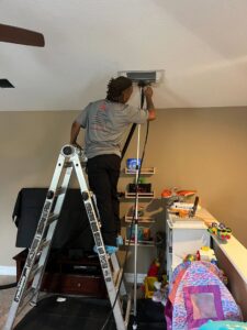 An HVAC technician on a ladder cleaning an indoor vent for Timely Air Conditioning in Riverview, FL.