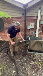 An HVAC technician cleaning an outdoor AC unit with a hose during maintenance by Radiant Heating & Cooling in Fishers, IN.