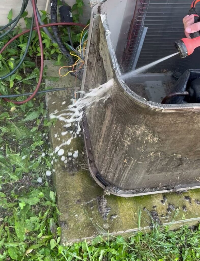 An HVAC technician cleaning an outdoor condenser unit with a hose for Direct Service HVAC & Maintenance in Lewisburg, OH.