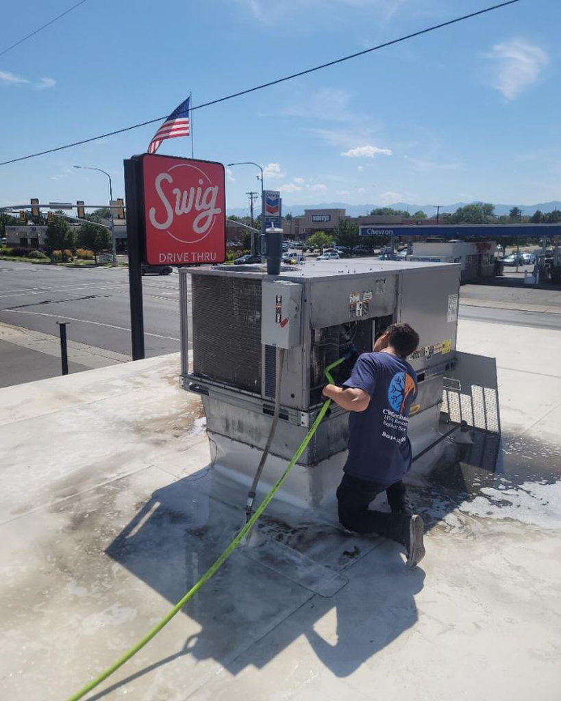 An HVAC technician cleaning a commercial rooftop unit with a hose for CW Mechanical HVACR in Lehi, UT.
