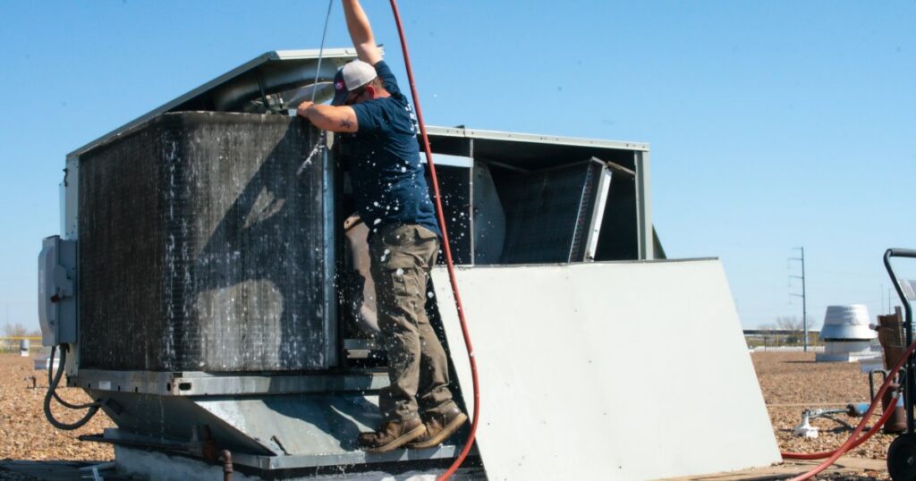 An HVAC technician from DMC Service, Inc. performing maintenance by cleaning a large commercial rooftop HVAC unit in Olathe, KS.