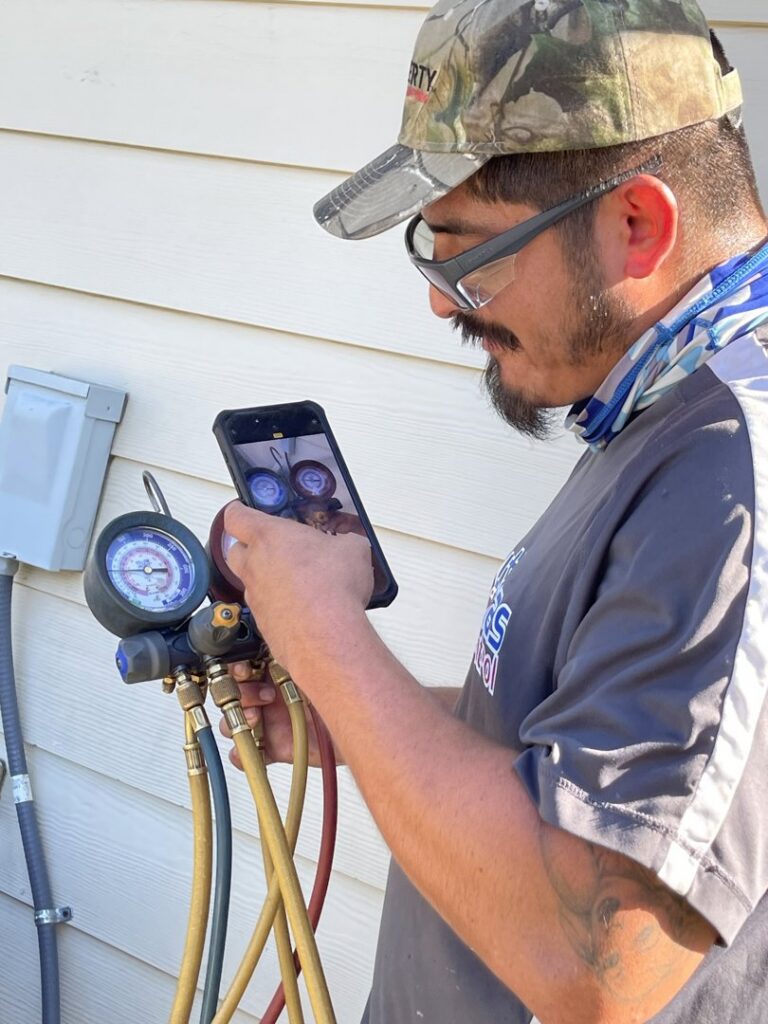 An HVAC technician documenting refrigerant gauge readings on an AC unit with a smartphone for South Texas Climate Control in San Antonio, TX