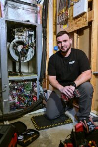 An Omega Air Solutions HVAC technician kneeling next to an open furnace unit, ready for repair in West Des Moines, IA.